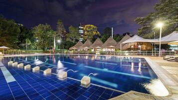 A swimming pool with a tiled deck, surrounded by thatched-roof structures and trees.