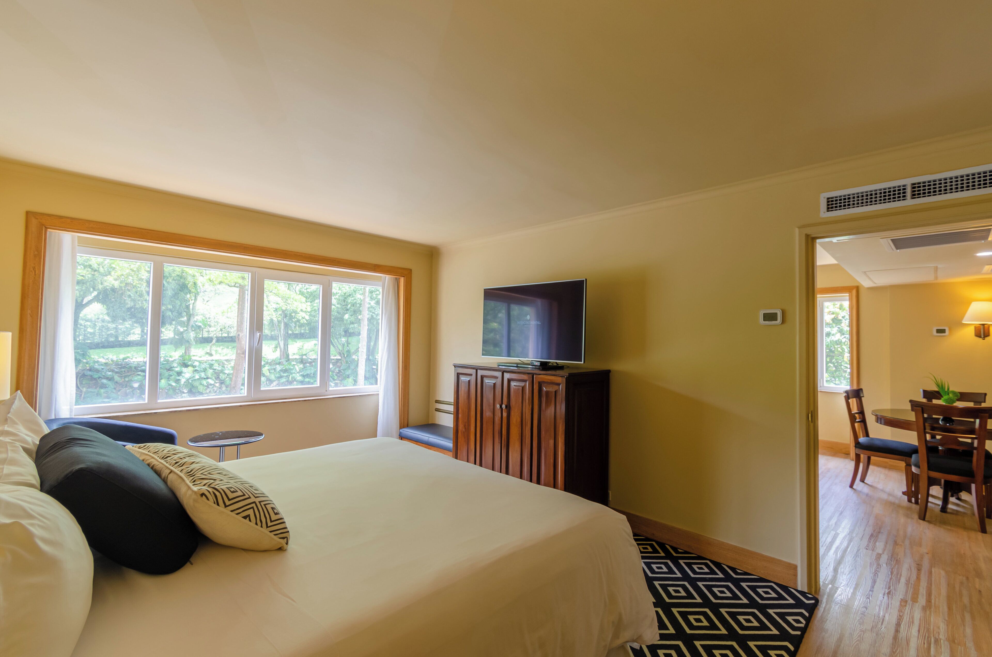 Quarto de hotel com uma cama, uma televisão sobre um móvel de madeira, um espaço de refeições com mesa e cadeiras, e uma janela com vista para o verde.