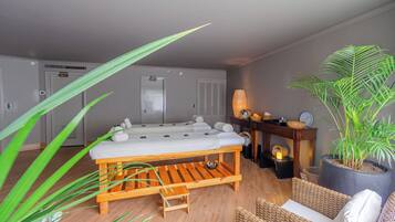 Une pièce comprenant une table de massage, un petit banc en bois, une chaise en osier, une plante en pot et une petite table d’appoint en bois avec une lampe.