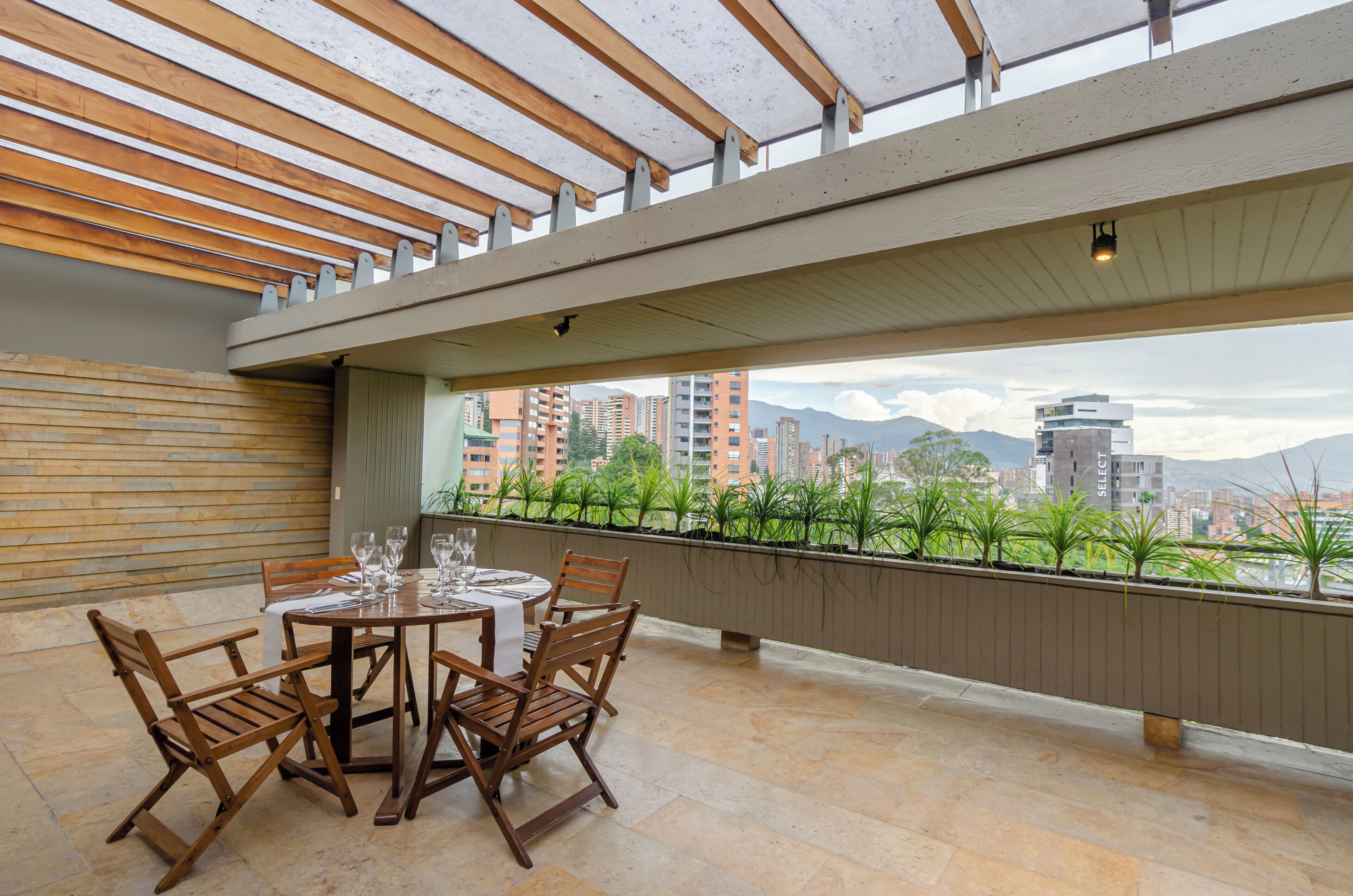 Terraza en la azotea con mesa y sillas de madera, vista a un paisaje urbano y barandilla adornada con plantas.
