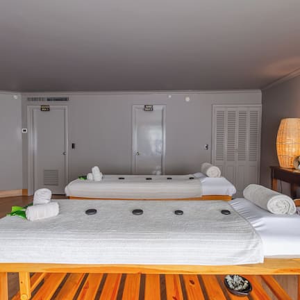 A room with two beds, a massage table, a wooden bench, and a small table with candles and a basket.
