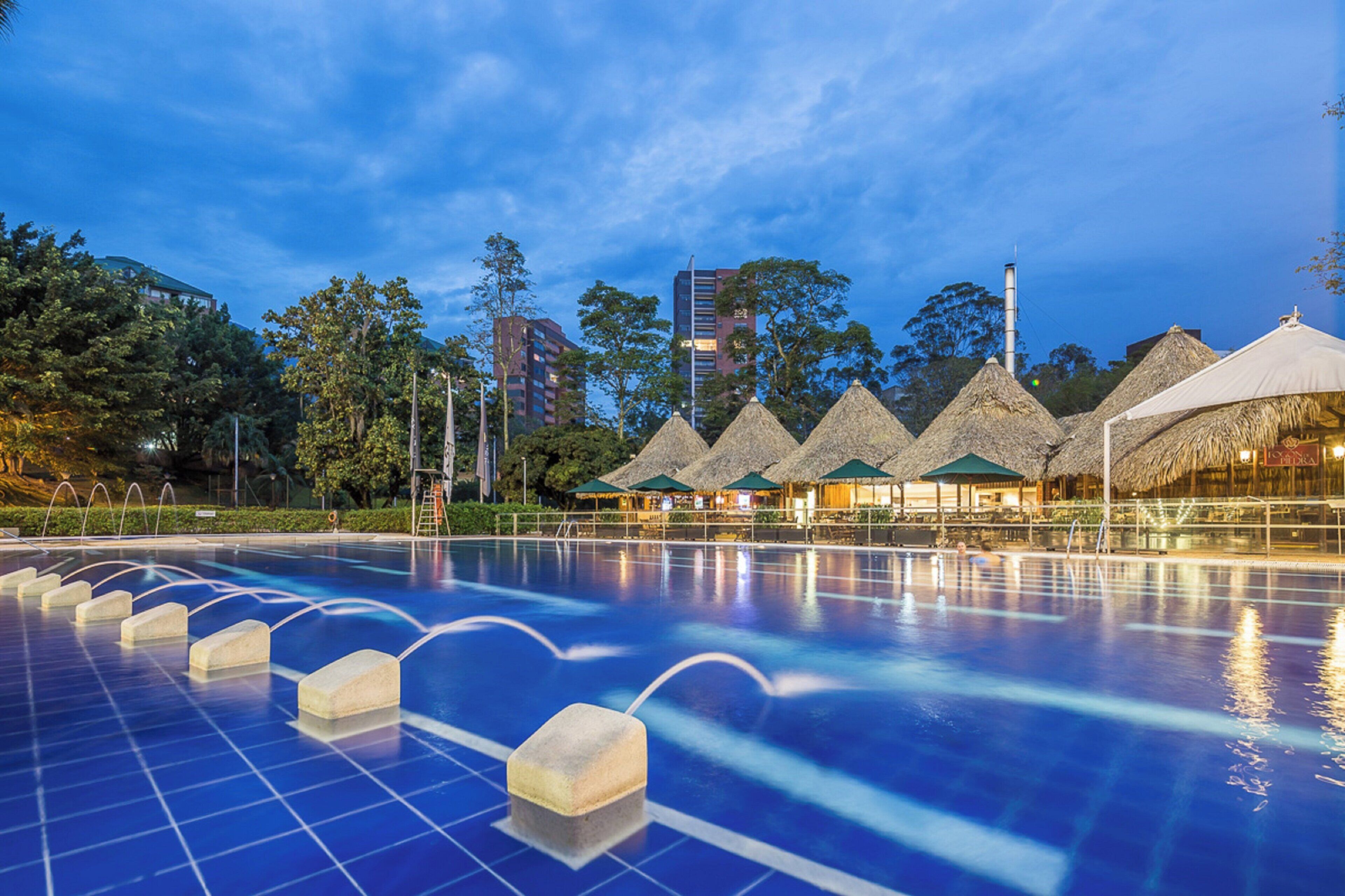 A swimming pool with a solar panel array in the foreground, surrounded by thatched-roof structures and modern buildings in the background.