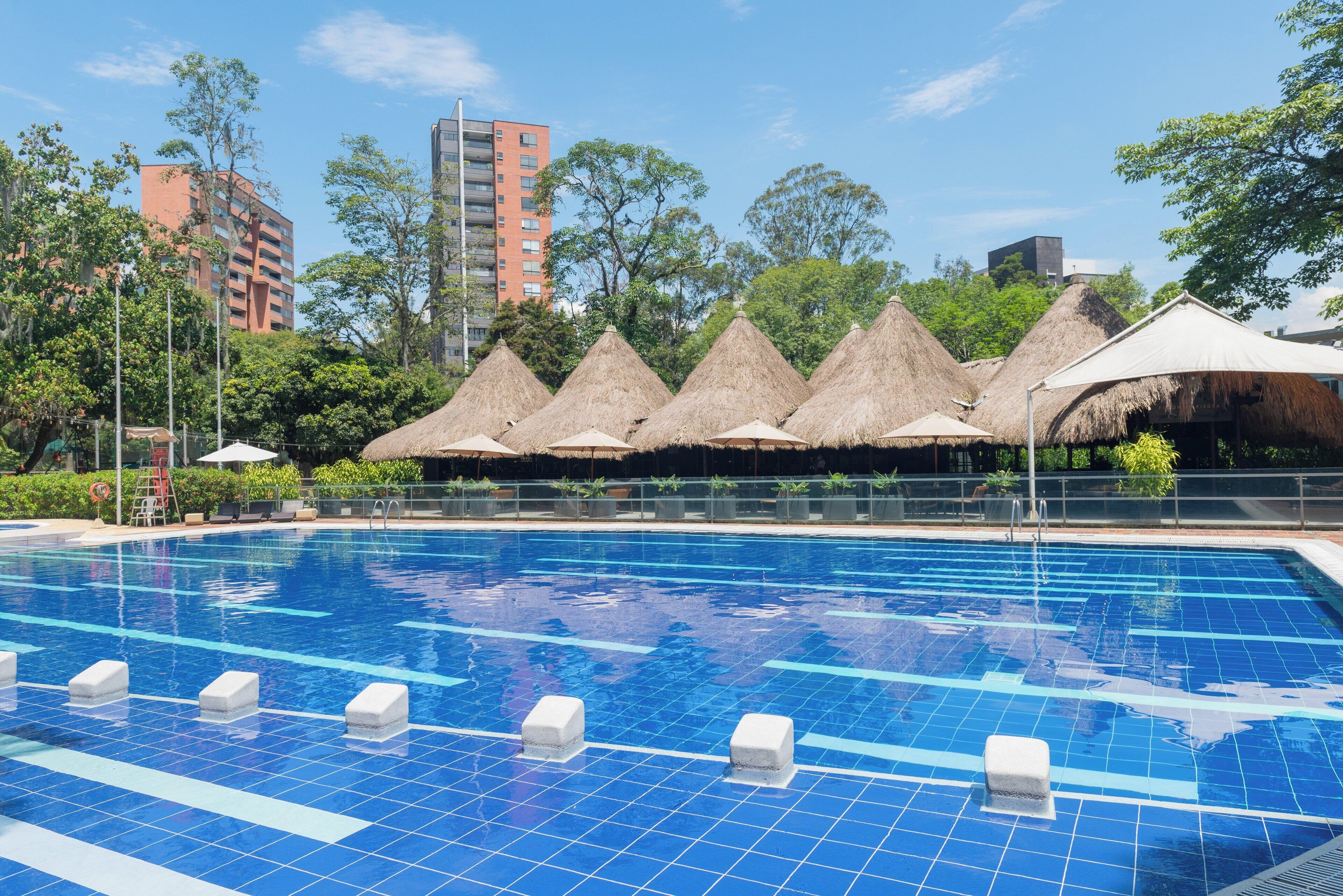 Una piscina con baldosas azules cristalinas, rodeada de sombrillas de paja y follaje.