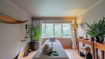 A spa room with a massage table, potted plants, a wooden shelf, and a window with a view of greenery.