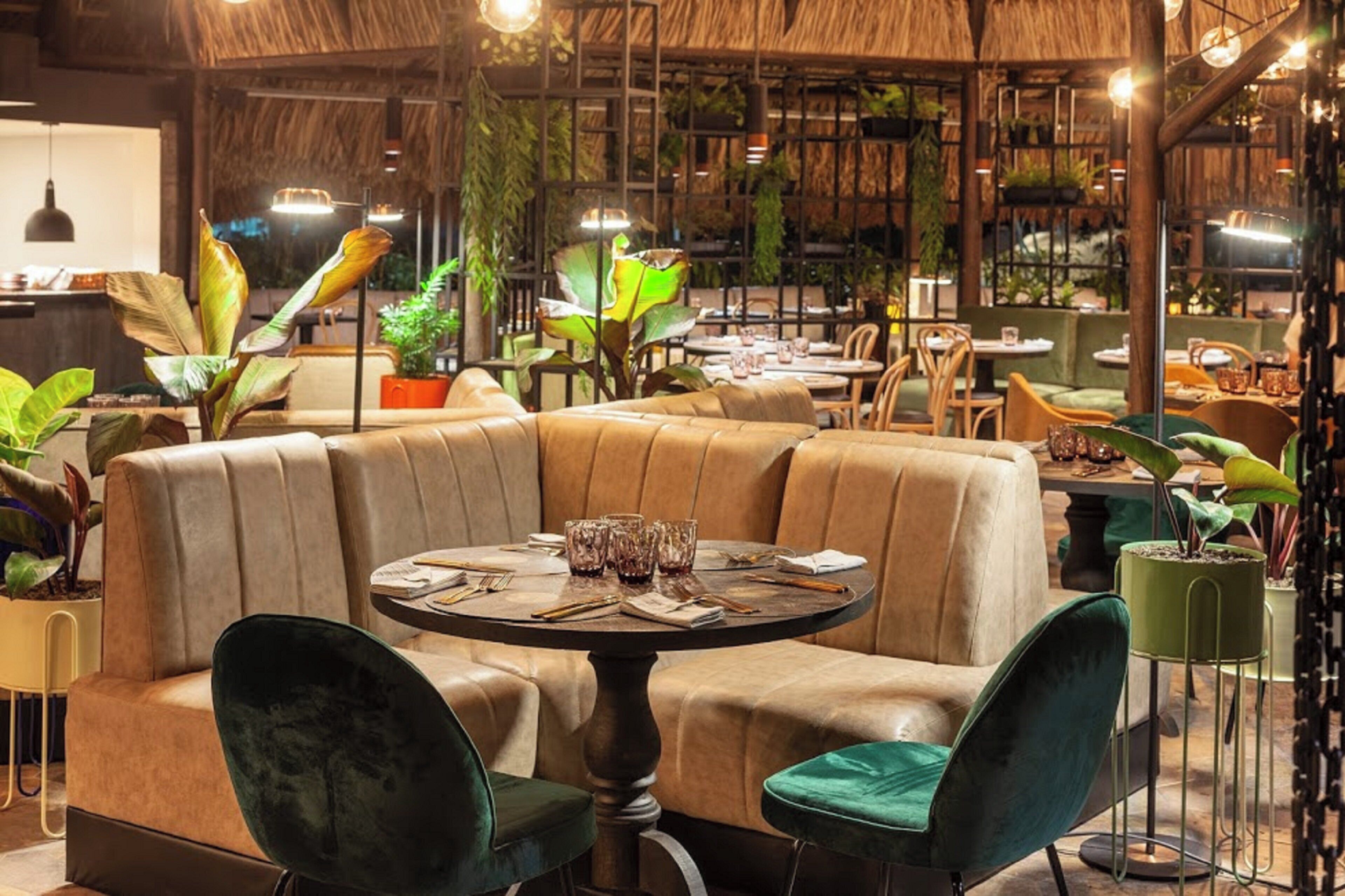 Interior de restaurante con mesas redondas, asientos tipo cabina y plantas en macetas.