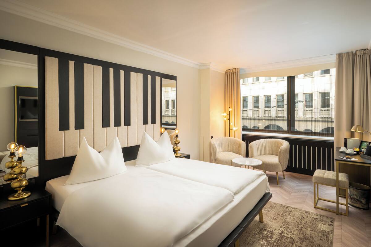 Ein Hotelzimmer mit einem großen Bett, einem Sessel, einem kleinen Tisch, einem Schreibtisch und Blick auf die Stadt.