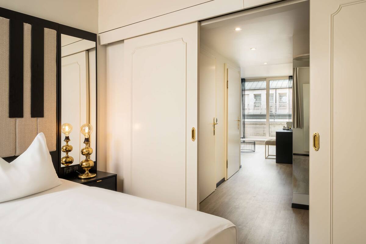 Ein Hotelzimmer mit Bett, Nachttisch, Lampe, Kleiderschrank und Blick auf die Stadt durch große Fenster.