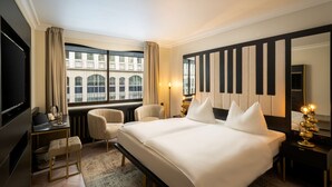 Habitación de hotel con una cama grande, un escritorio con una computadora portátil, una silla, una mesita de noche con una lámpara y vista a un edificio a través de la ventana.