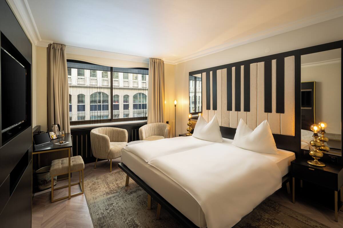 Ein Hotelzimmer mit einem großen Bett, einem Schreibtisch, einem Sessel, einem Fernseher und Blick auf ein Gebäude durch das Fenster.