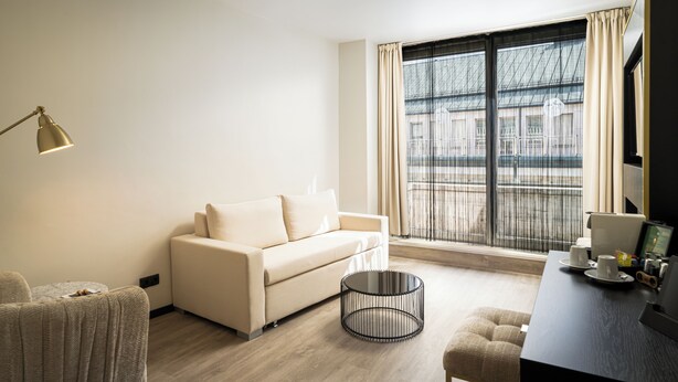 A modern hotel room with a sofa, a round coffee table, a desk with a computer, and a large window with curtains. - elaya hotel vienna city center, Trademark by Wyndham (Vienna)