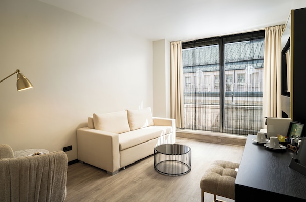 Una habitación de hotel moderna con un sofá, una mesita redonda, un escritorio con una computadora y un gran ventanal con cortinas.