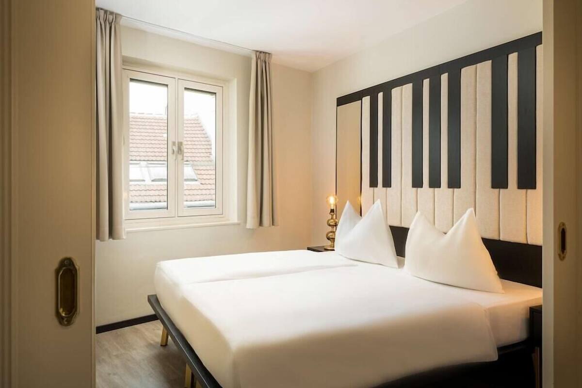 Ein Hotelzimmer mit zwei Betten, einem Fenster mit Vorhängen, einer Wandleuchte und Blick auf Gebäude.
