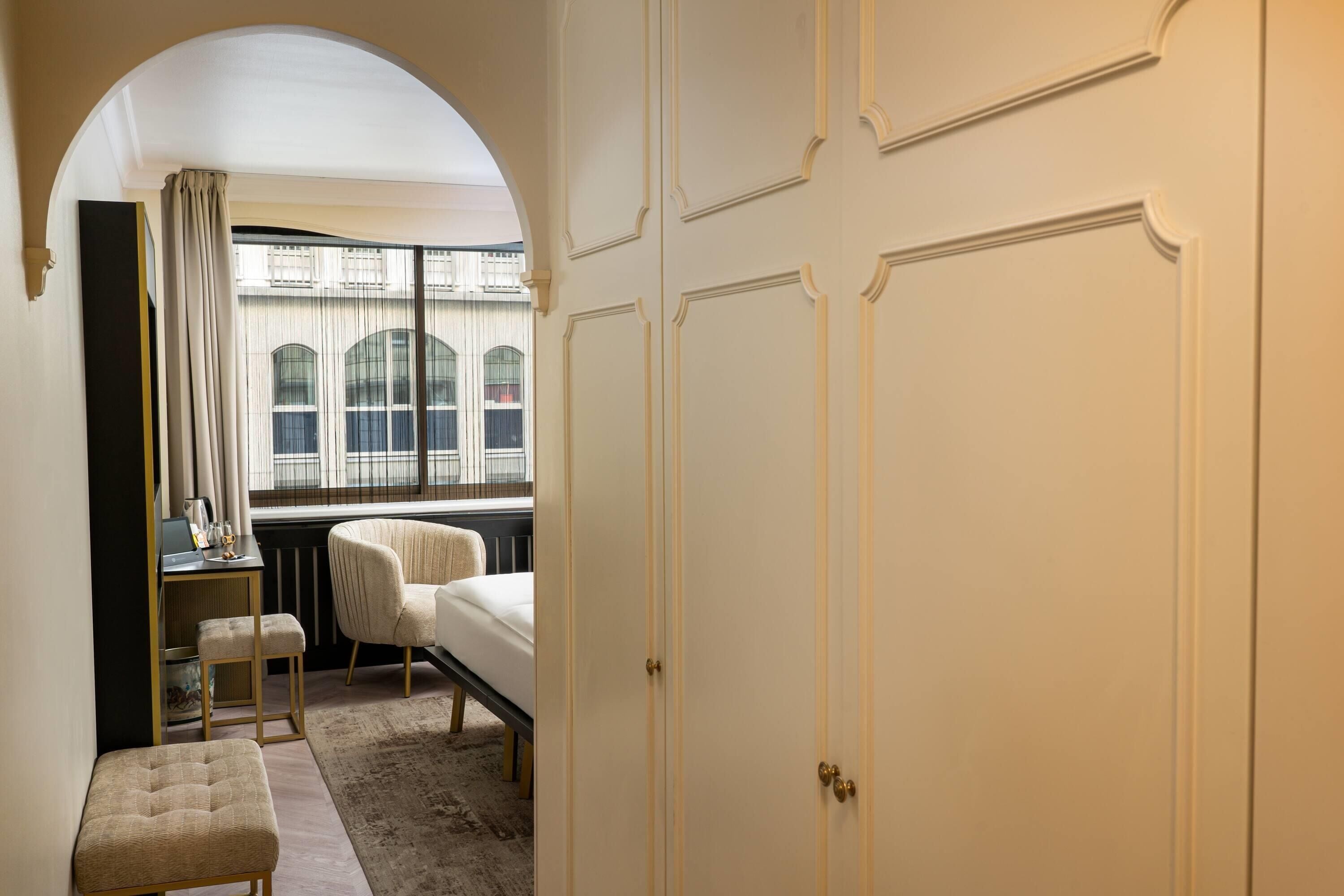 Ein Hotelzimmer mit einem Bett, einem Schreibtisch mit Stuhl, einem kleinen Hocker und einem Fenster mit Vorhängen.