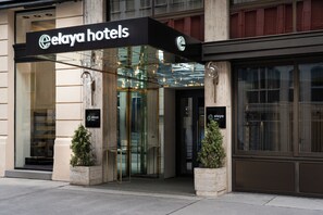 El exterior del hotel Elaya, con acceso de vidrio, un cartel sobre la puerta y dos plantas en macetas a cada lado.