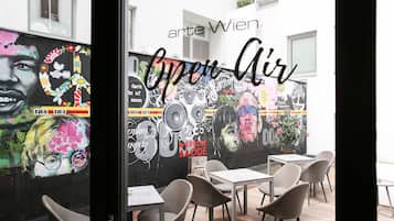 Un área de café al aire libre con mesas y sillas, un mural en la pared y el texto "arte Wien Open Air".