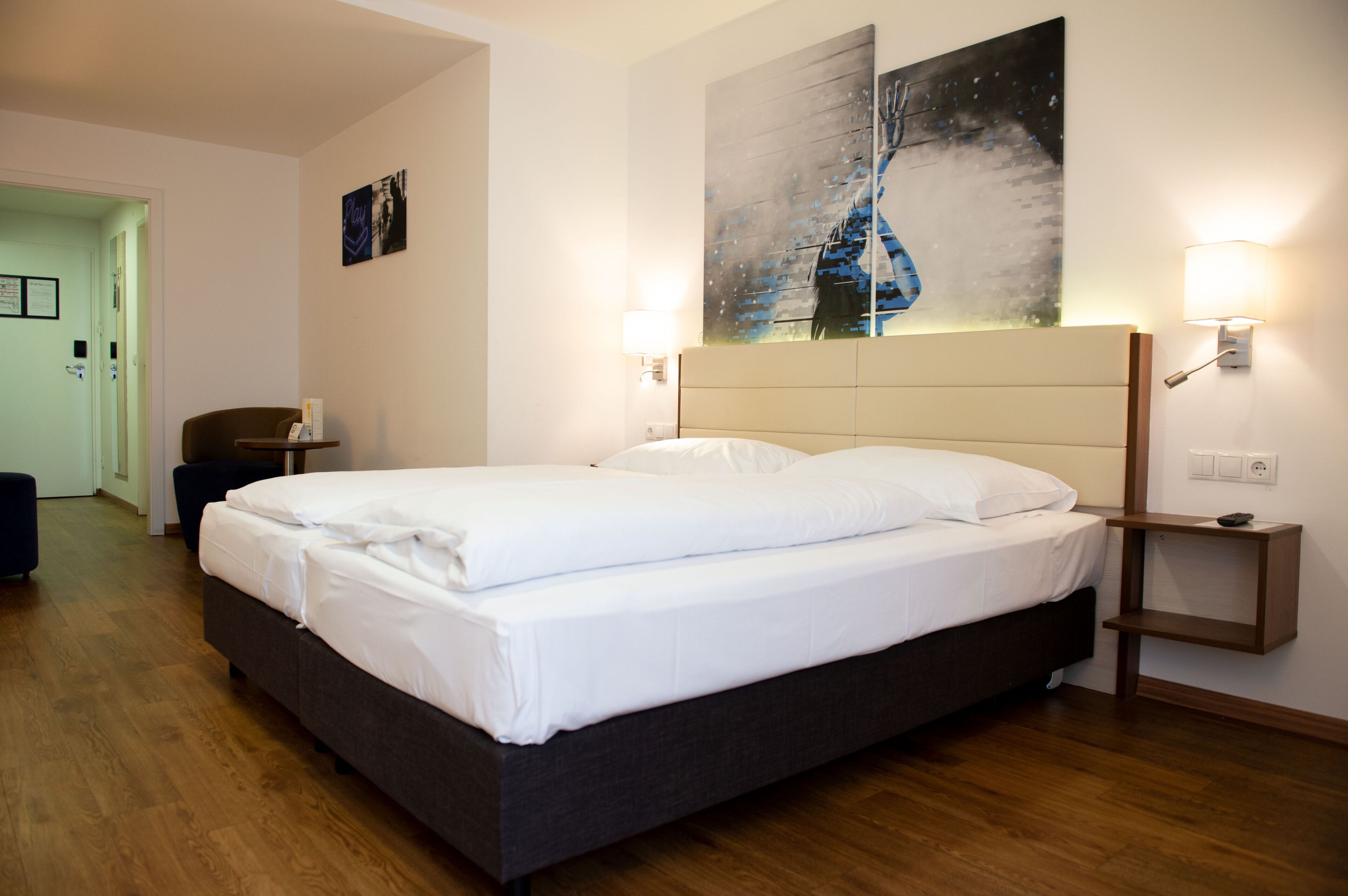 A hotel room with a large bed, bedside lamps, a headboard, a wall-mounted artwork, and a door leading to another room.