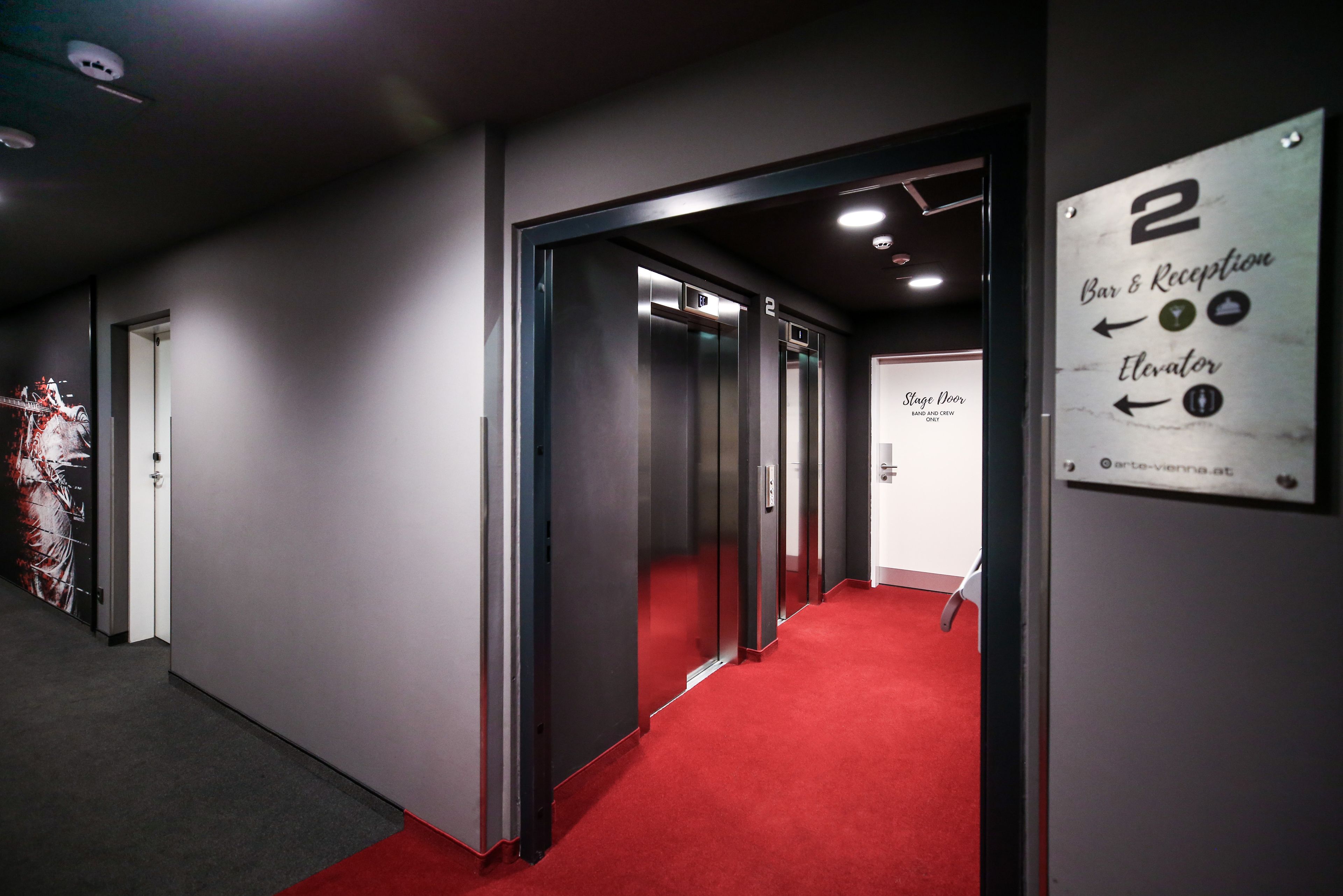 Un pasillo con alfombra roja que conduce a un ascensor, un letrero de bar y recepción, y una puerta de acceso al escenario.