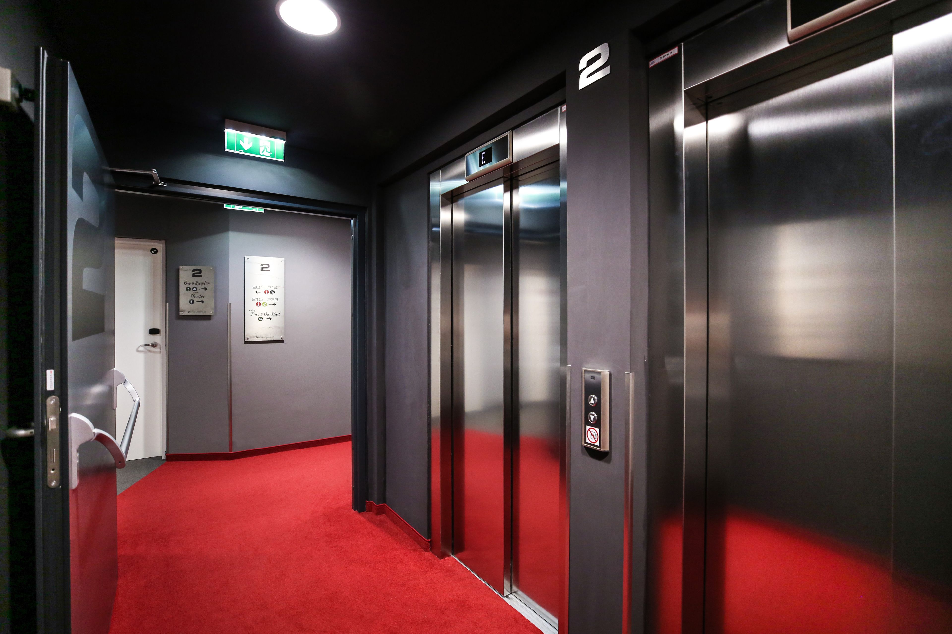Un ascensor con piso alfombrado en rojo, señal de salida de emergencia y una puerta con el número 2.