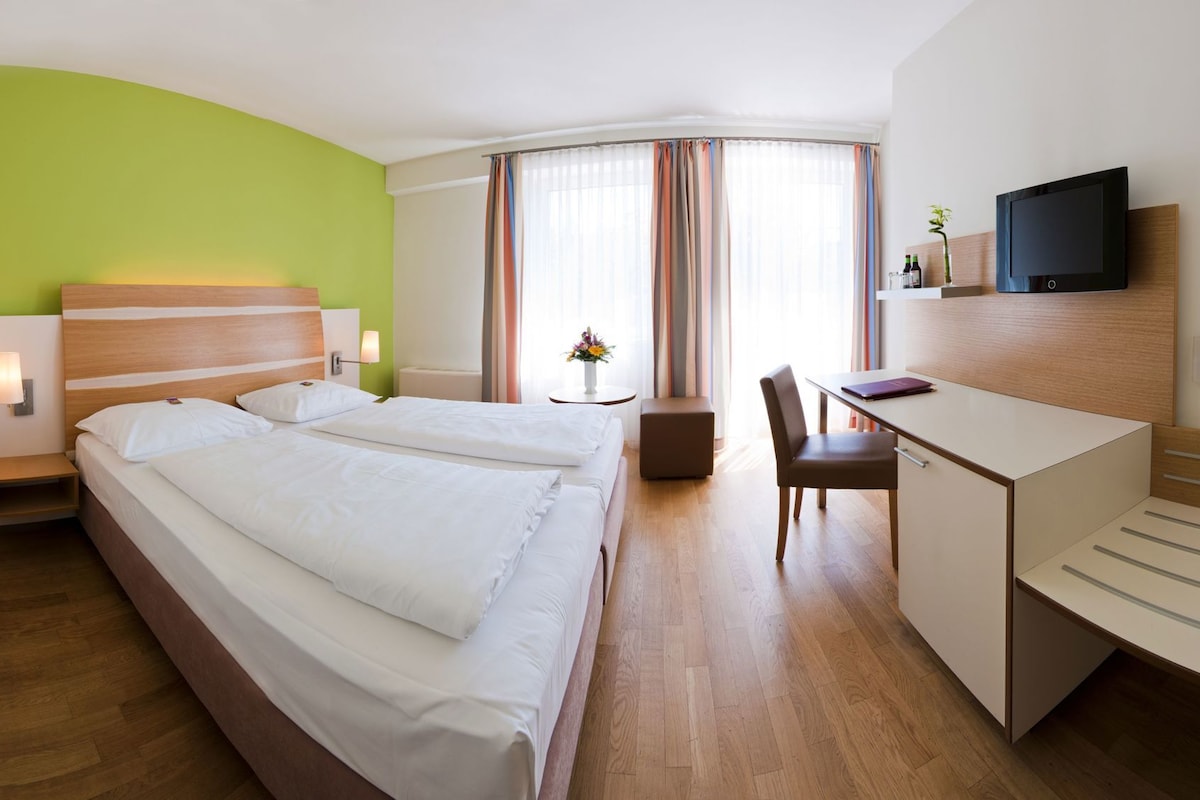 A hotel room with two beds, a desk, a chair, a TV, and a small table with a flower vase.