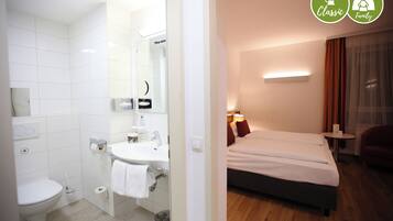 Quarto de hotel com banheiro, uma cama, mesa de cabeceira e um pequeno espaço para sentar.