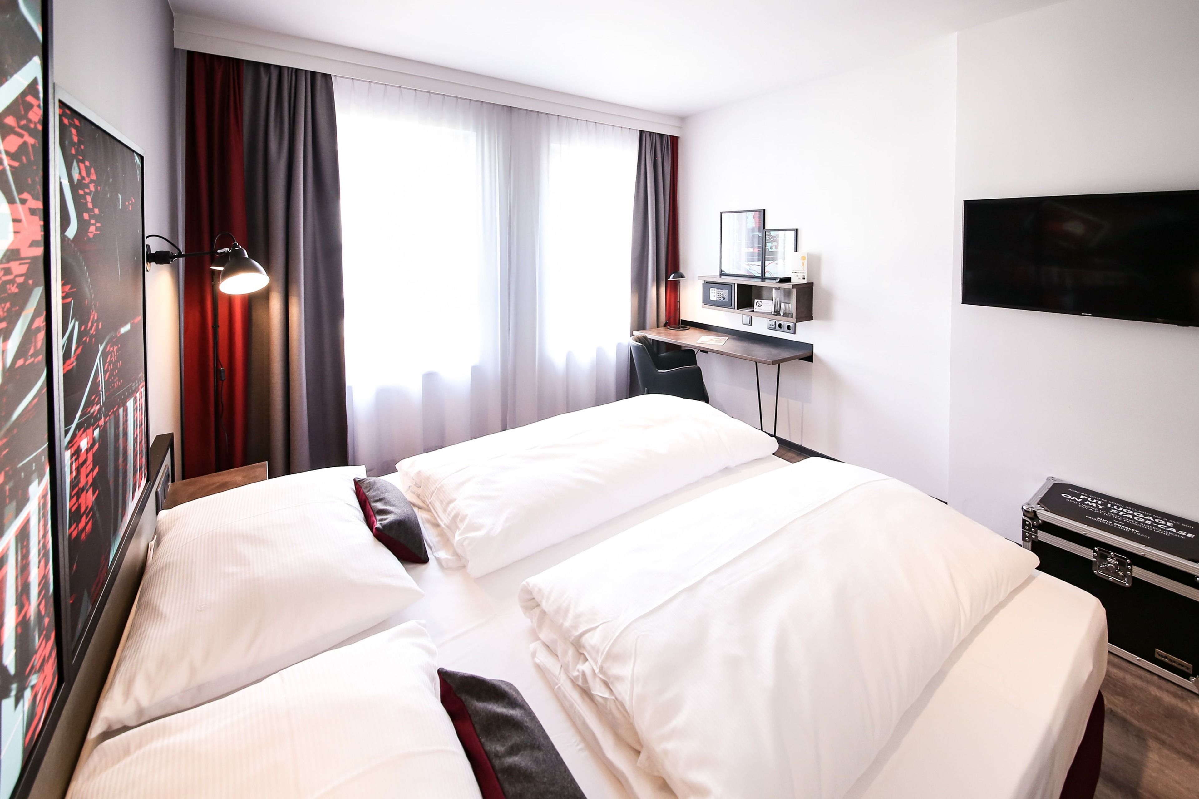 A hotel room with a large bed, a desk with a computer, a flat-screen TV, and a large window with curtains.