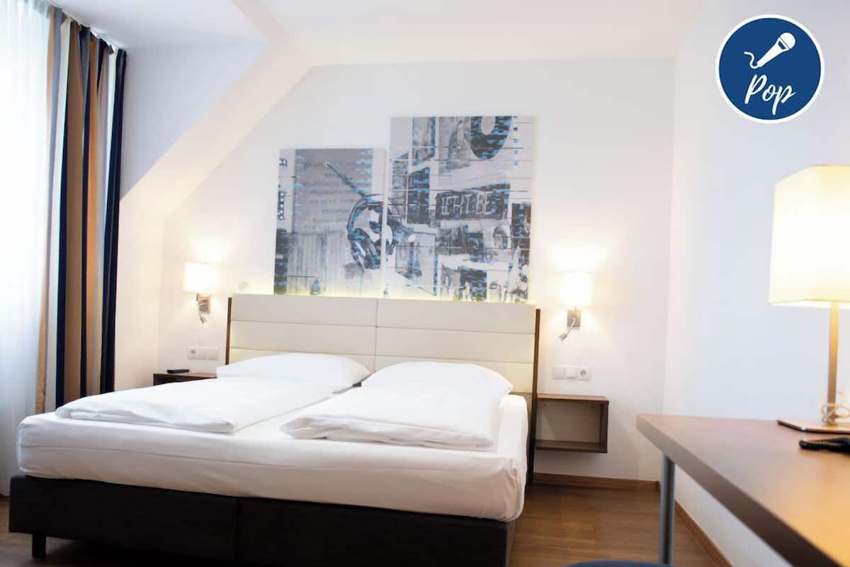 Une chambre d’hôtel avec un lit, deux tables de chevet, un bureau et une lampe. Un grand tableau abstrait orne le mur au-dessus du lit.