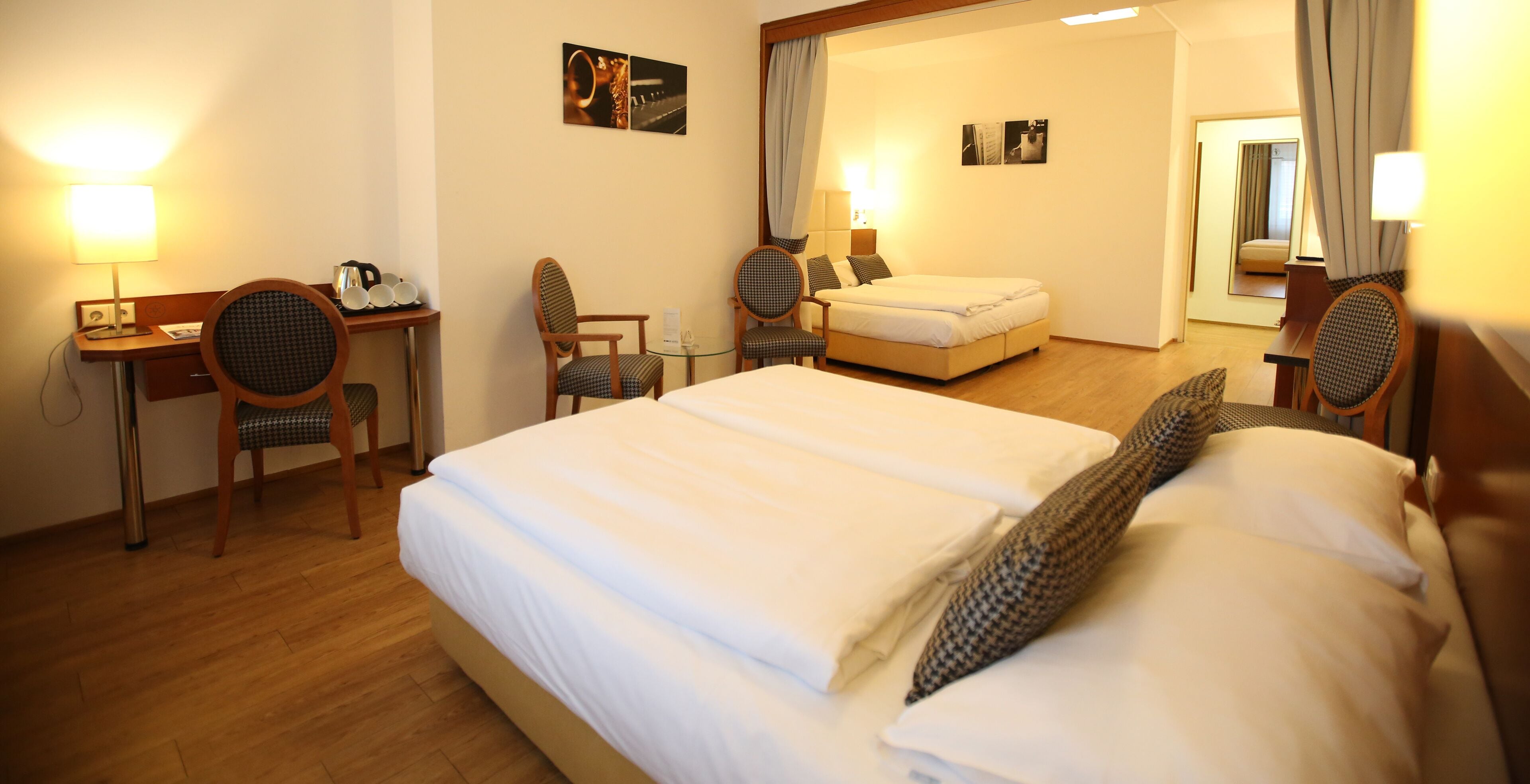 Quarto de hotel com uma cama grande, uma escrivaninha com luminária, duas cadeiras e uma mesa pequena.