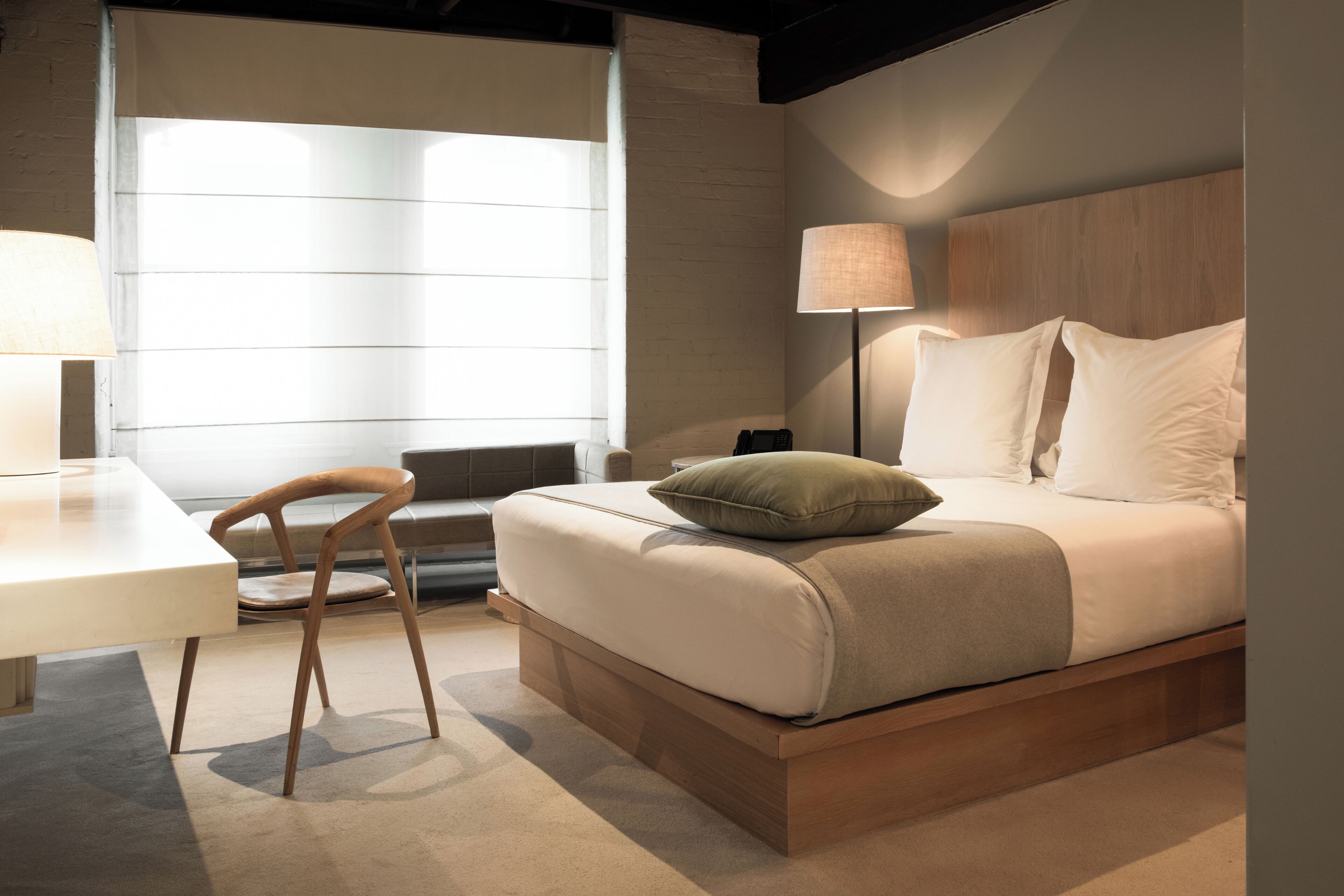 A modern hotel room with a bed, bedside table, lamp, and a view of the outdoors.