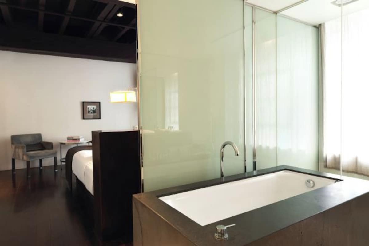 A modern hotel room with a large bathtub, a bed, a small table with a chair, and a framed picture on the wall.