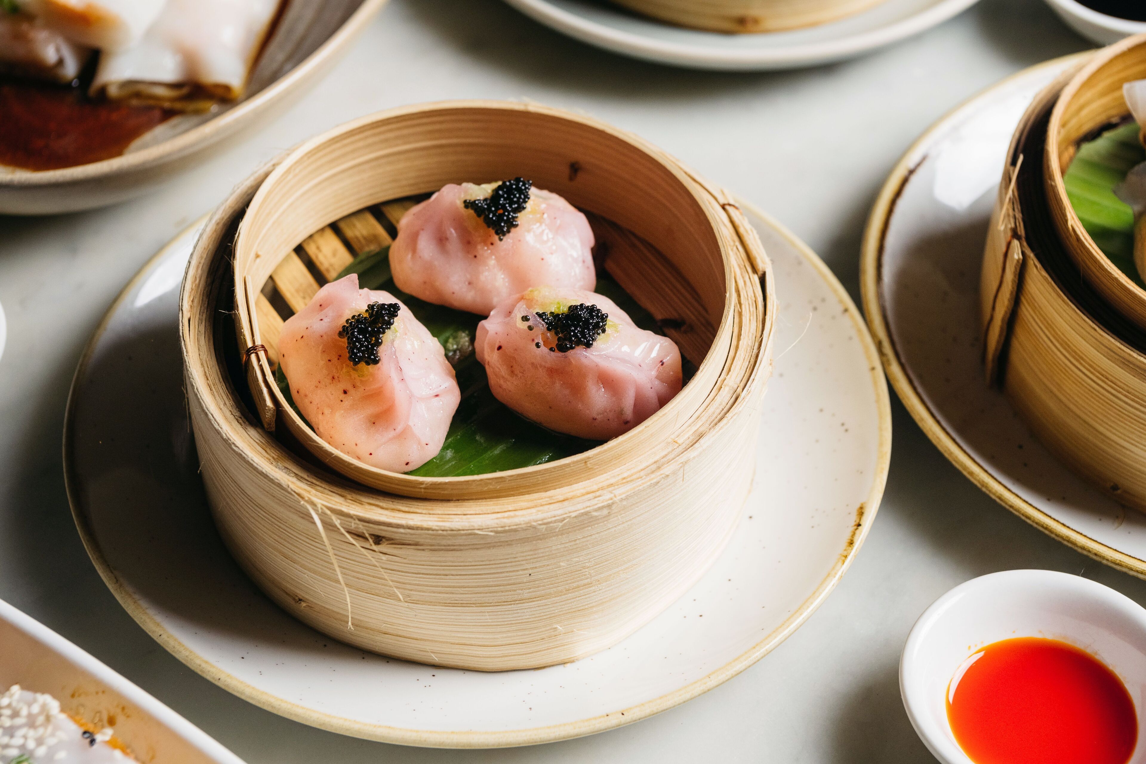 A table with a bamboo steamer containing three dumplings, a small bowl of red sauce, and a plate with a white bowl.