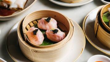A table with a bamboo steamer containing three dumplings, a small bowl of red sauce, and a plate with a white bowl.