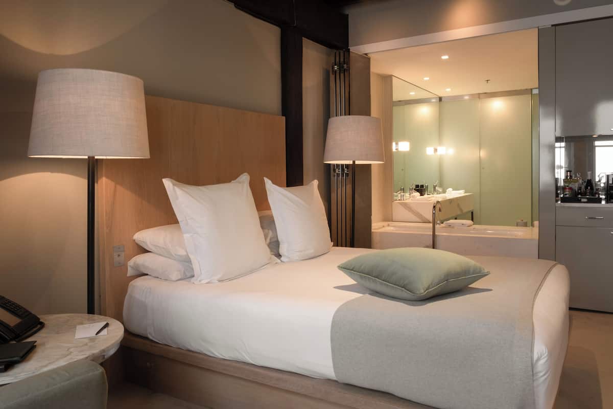 A modern hotel room with a neatly made bed, bedside lamps, a telephone, and a bathroom visible through an open door.
