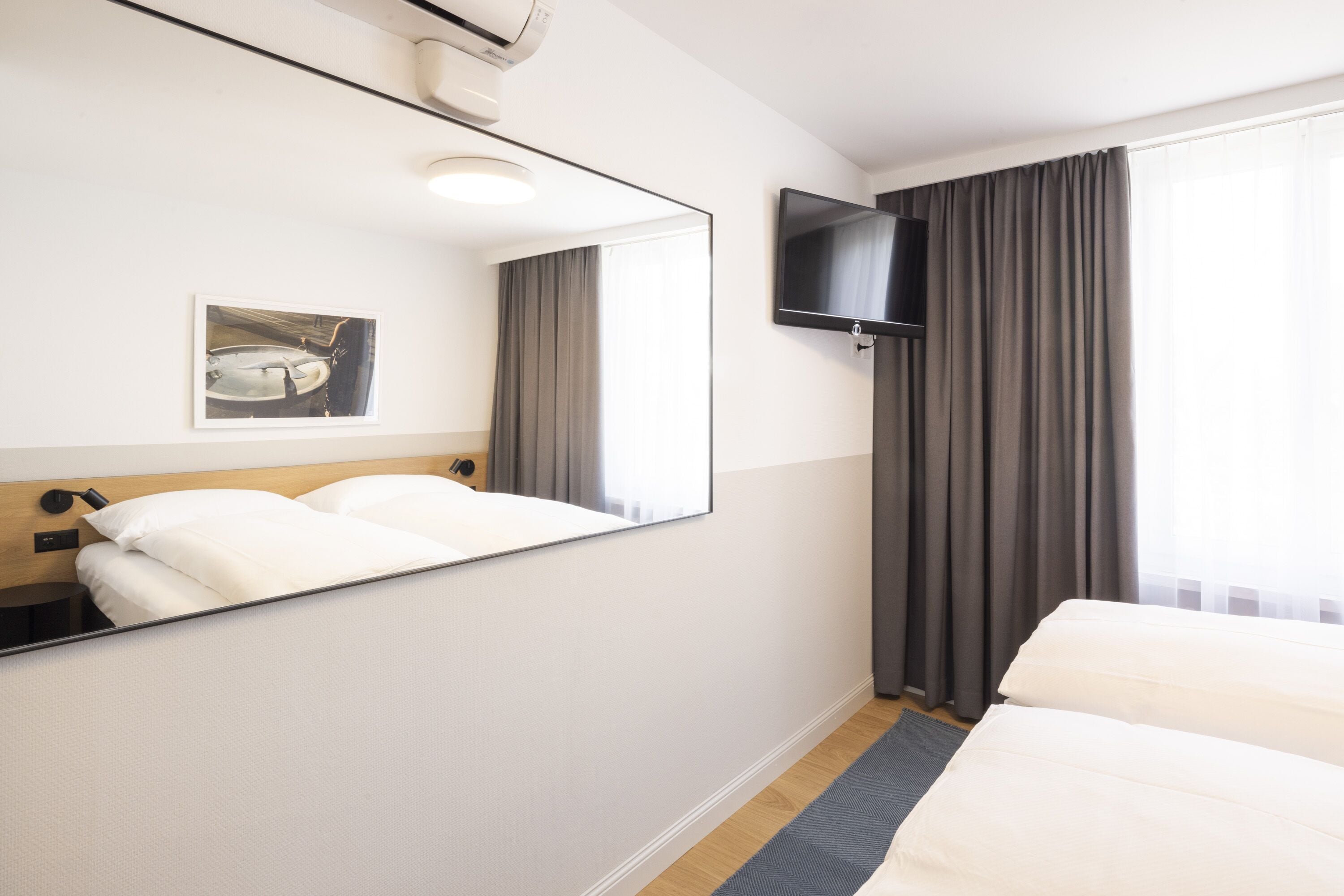 A hotel room with a large bed, a bedside table, a mirror, a flat-screen TV mounted on the wall, and a window with curtains.