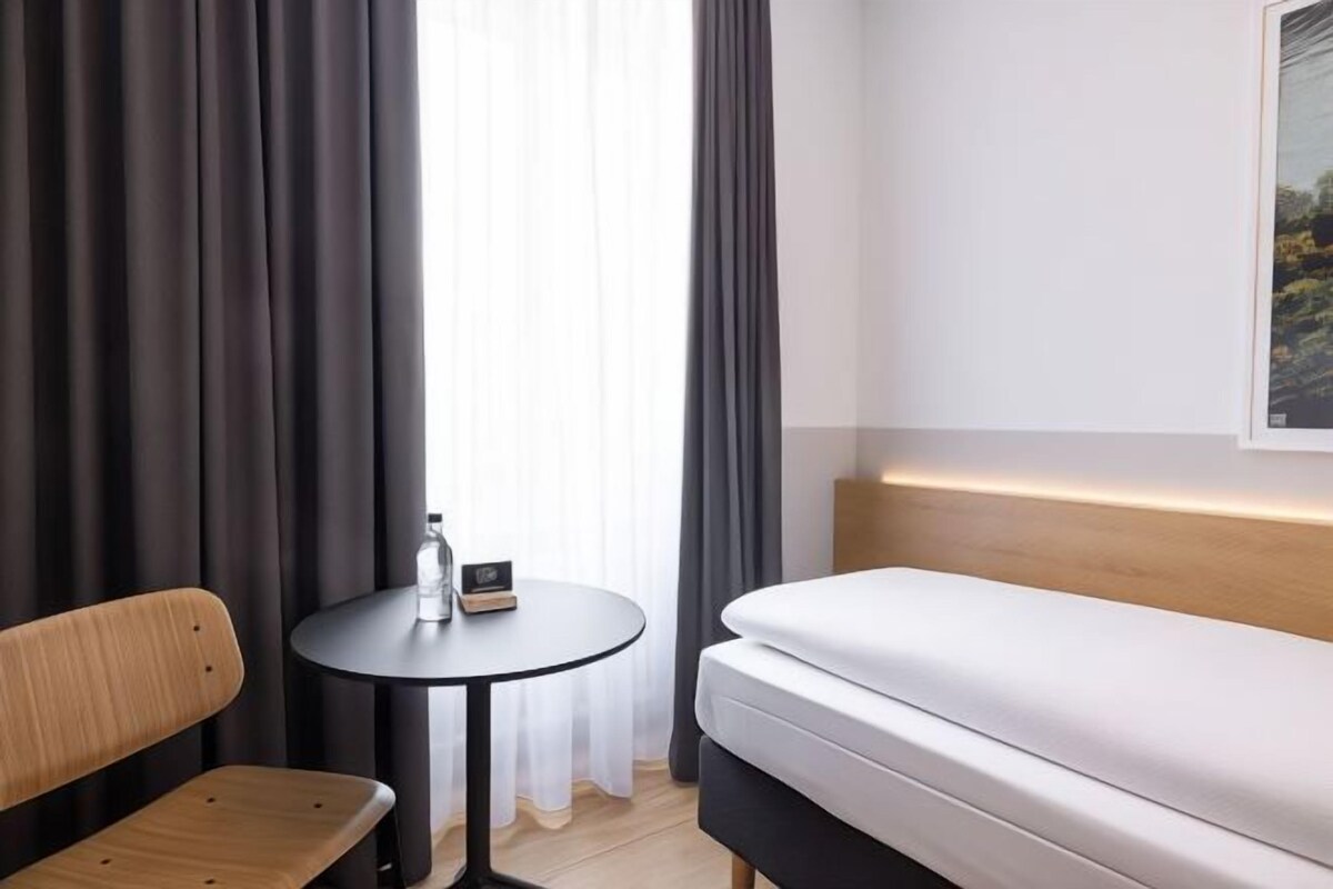 A hotel room with a bed, a wooden chair, a small round table with a bottle and a box, and a window with curtains.