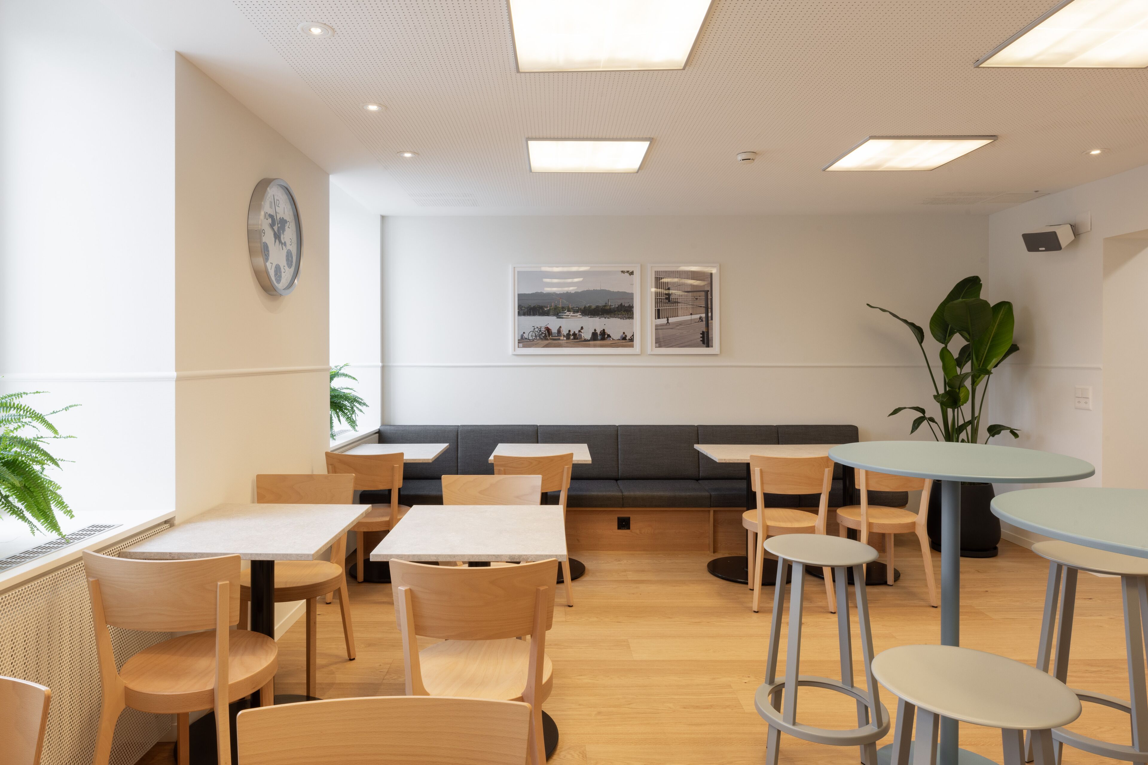 Un café moderne avec des tables et des chaises en bois, un coin salon avec un canapé et une grande fenêtre donnant sur l’extérieur.