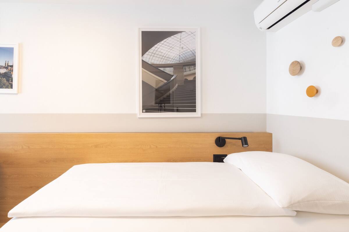 Ein Hotelzimmer mit einem hölzernen Kopfteil, einem Bett mit weißer Bettwäsche, einem gerahmten Bild eines Gebäudes und einer wandmontierten Klimaanlage.
