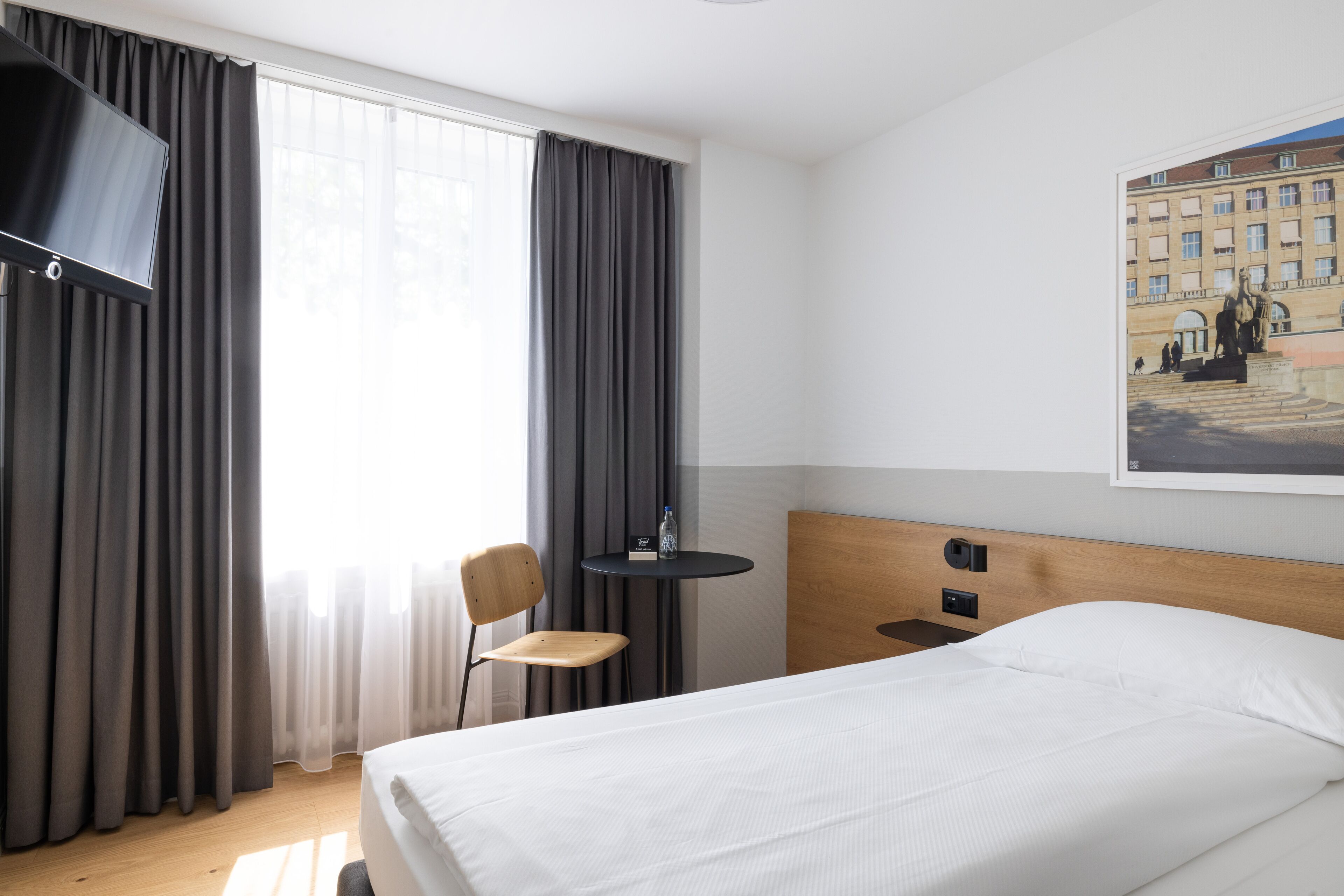 A hotel room with a bed, a chair, a small table, a TV, and a painting on the wall.