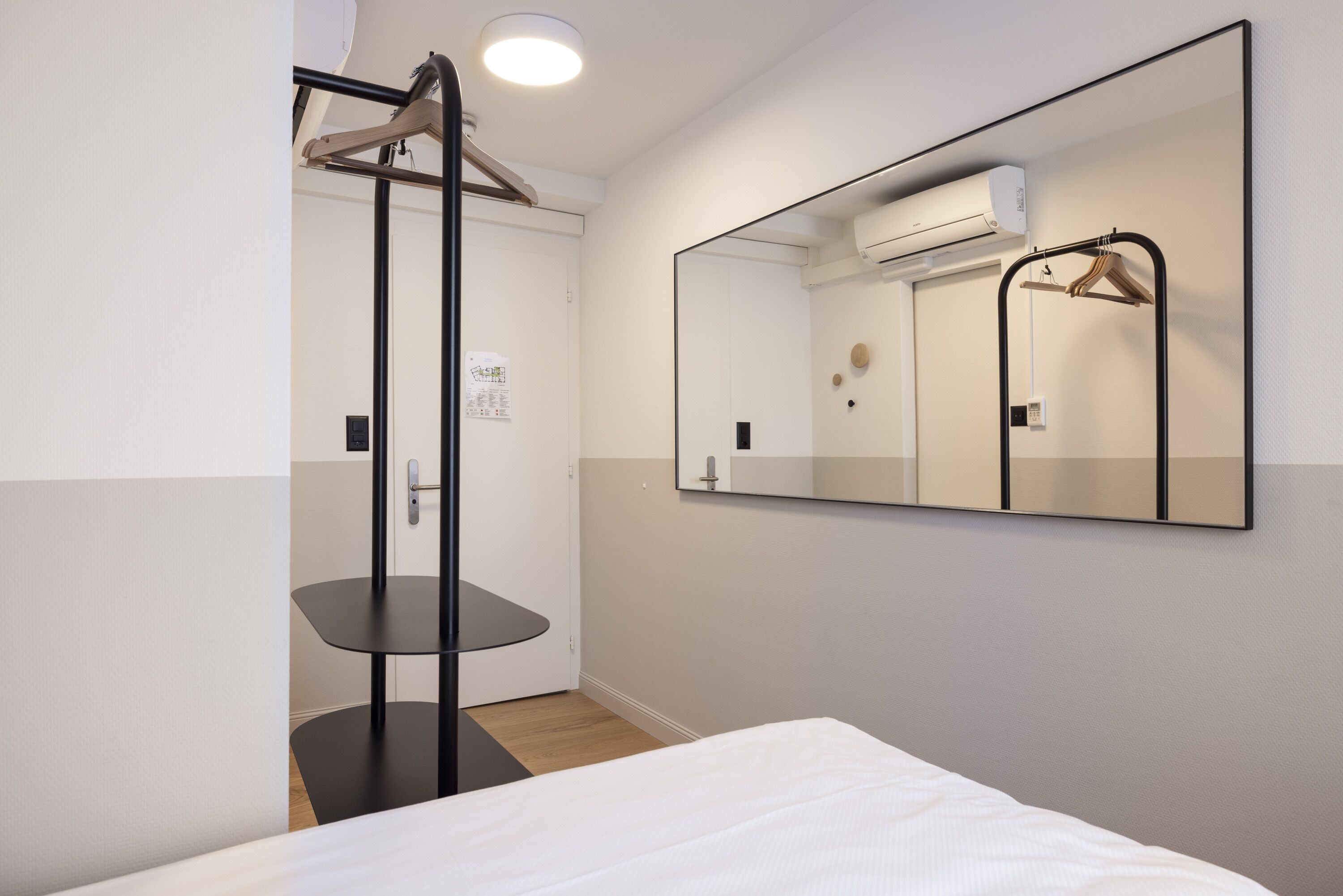 Ett hotellrum med en säng, ett litet bord, en spegel och en klädstång.