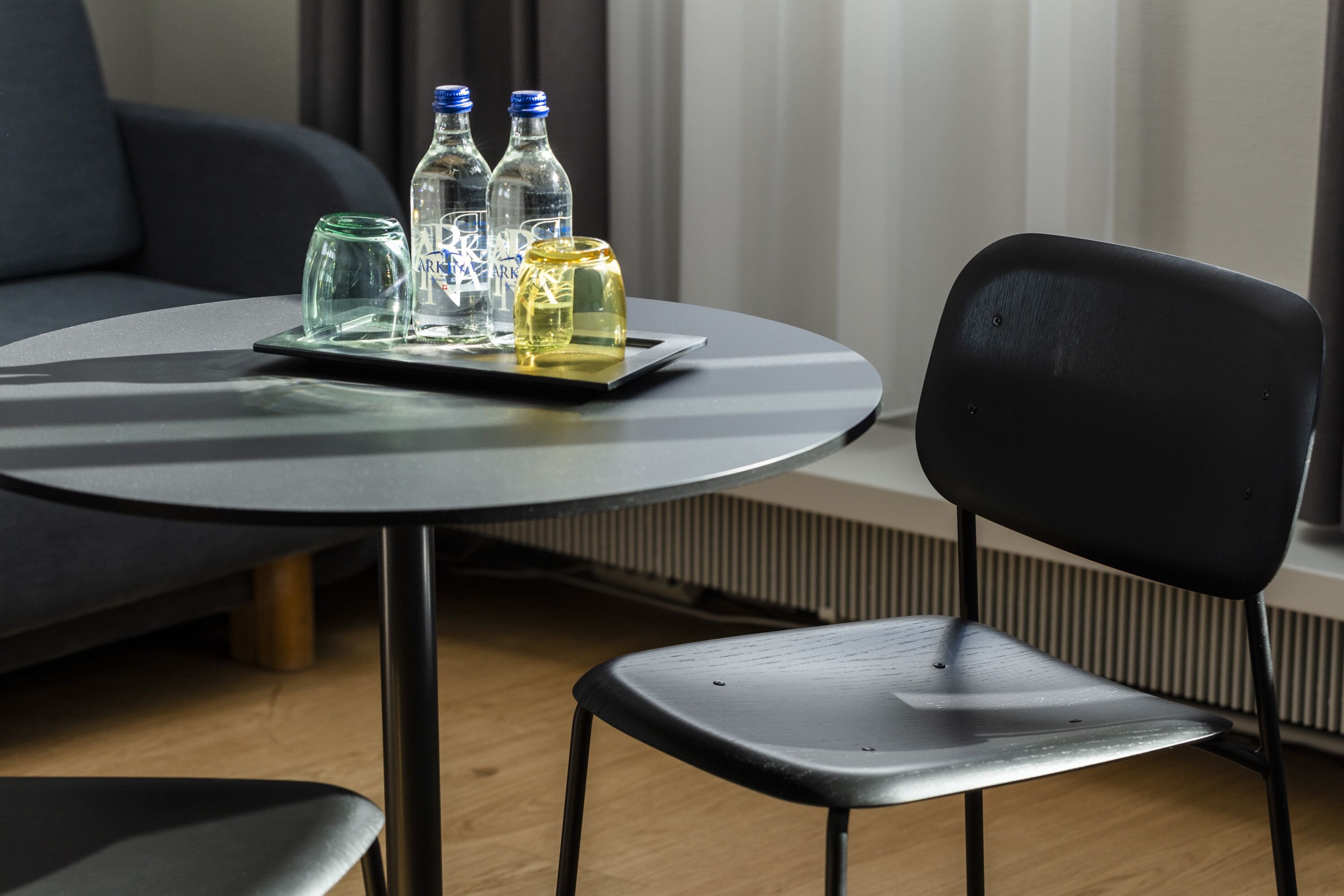 Una mesa redonda con una bandeja que sostiene tres botellas y un vaso, una silla negra y un sofá gris en una habitación con ventana y persianas.