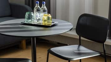 Una mesa redonda con una bandeja que sostiene tres botellas y un vaso, una silla negra y un sofá gris en una habitación con ventana y persianas.
