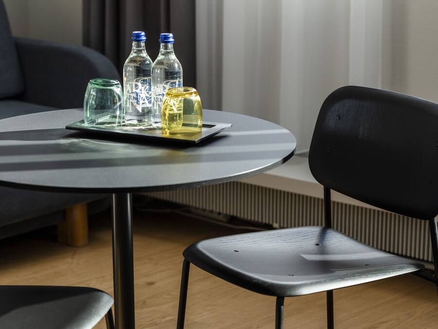 Una mesa redonda con una bandeja que sostiene tres botellas y un vaso, una silla negra y un sofá gris en una habitación con ventana y persianas.