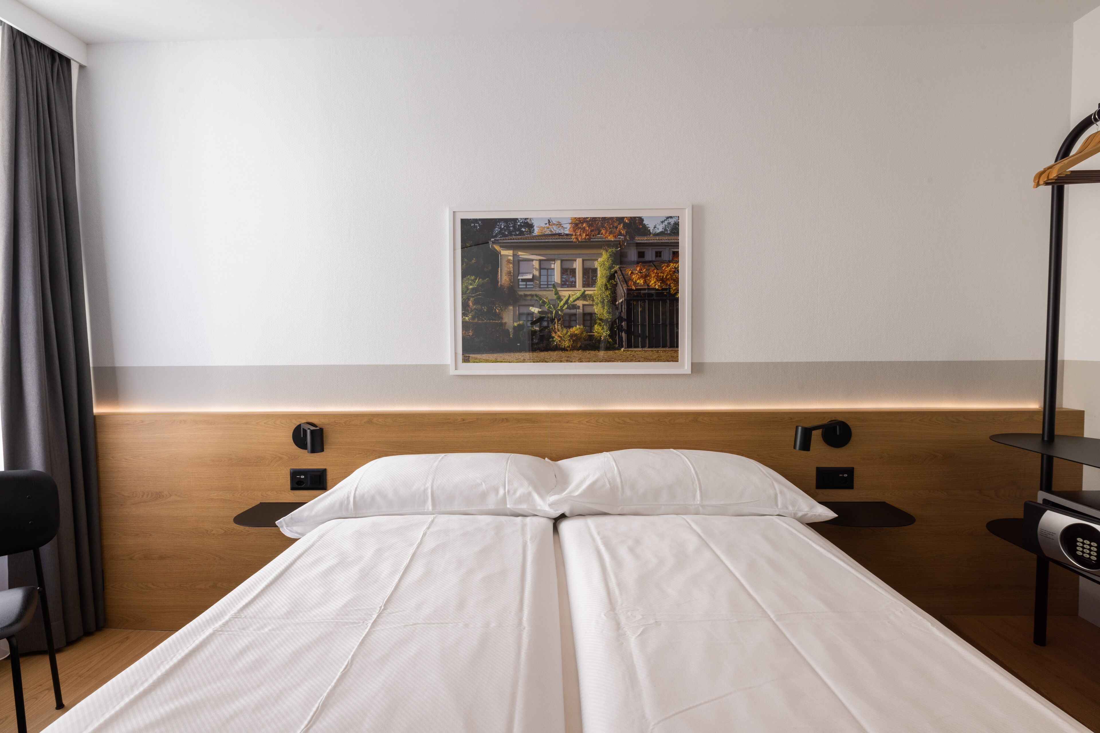A modern hotel room with a wooden headboard, a single bed with white linens, a nightstand with a lamp, and a framed picture on the wall.