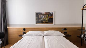 A modern hotel room with a wooden headboard, a single bed with white linens, a nightstand with a lamp, and a framed picture on the wall.