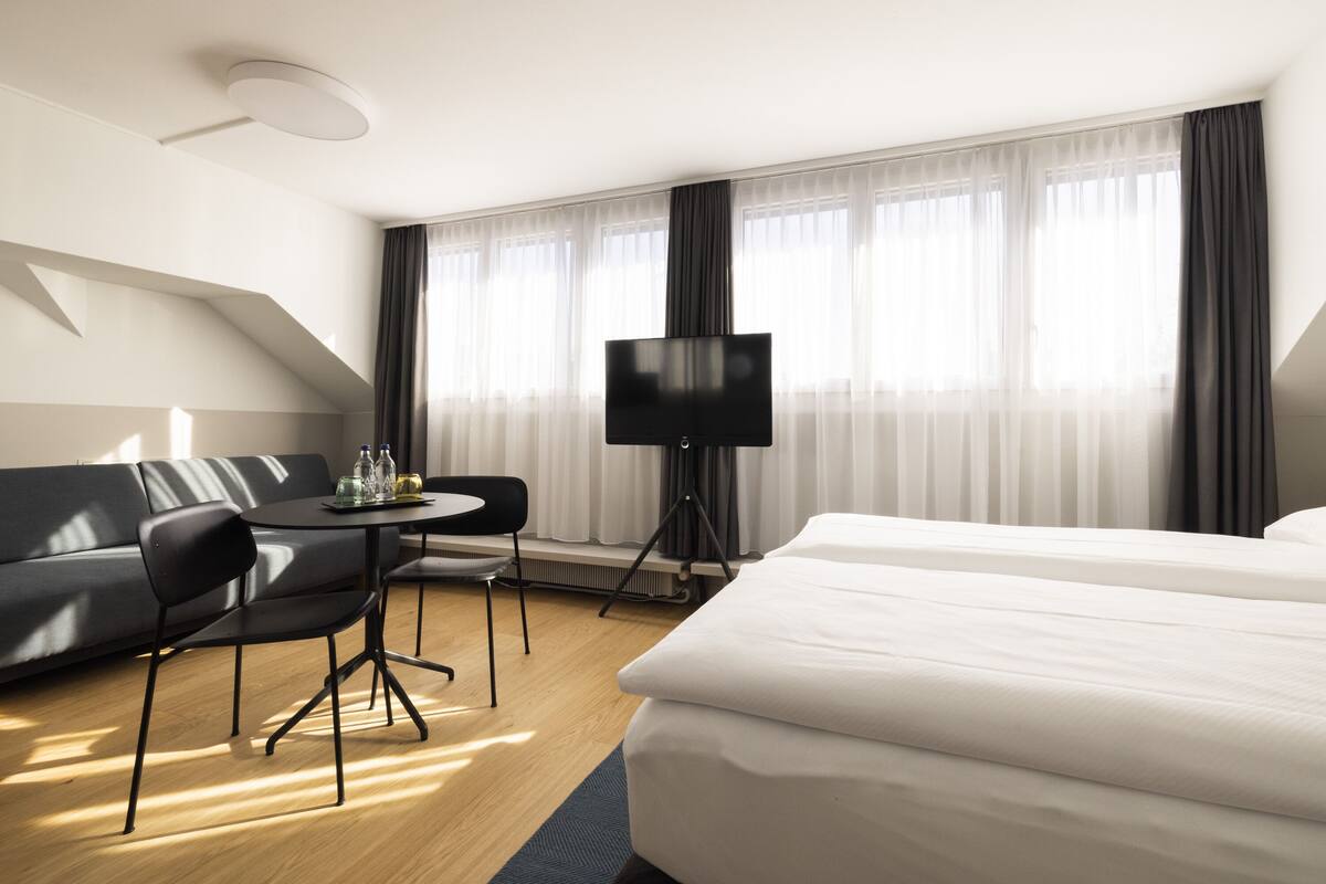 Ein modernes Hotelzimmer mit einem großen Bett, einer Sitzecke, einem Esstisch mit Stühlen, einem Flachbildfernseher und großen Fenstern mit durchsichtigen Vorhängen.