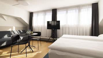 A modern hotel room with a large bed, a sofa, a dining table with chairs, a flat-screen TV, and large windows with sheer curtains.