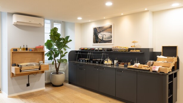 A modern kitchen with a central island, wooden shelves, a potted plant, and a variety of food items displayed on the counter. - Fred Hotel Hauptbahnhof (Zürich)
