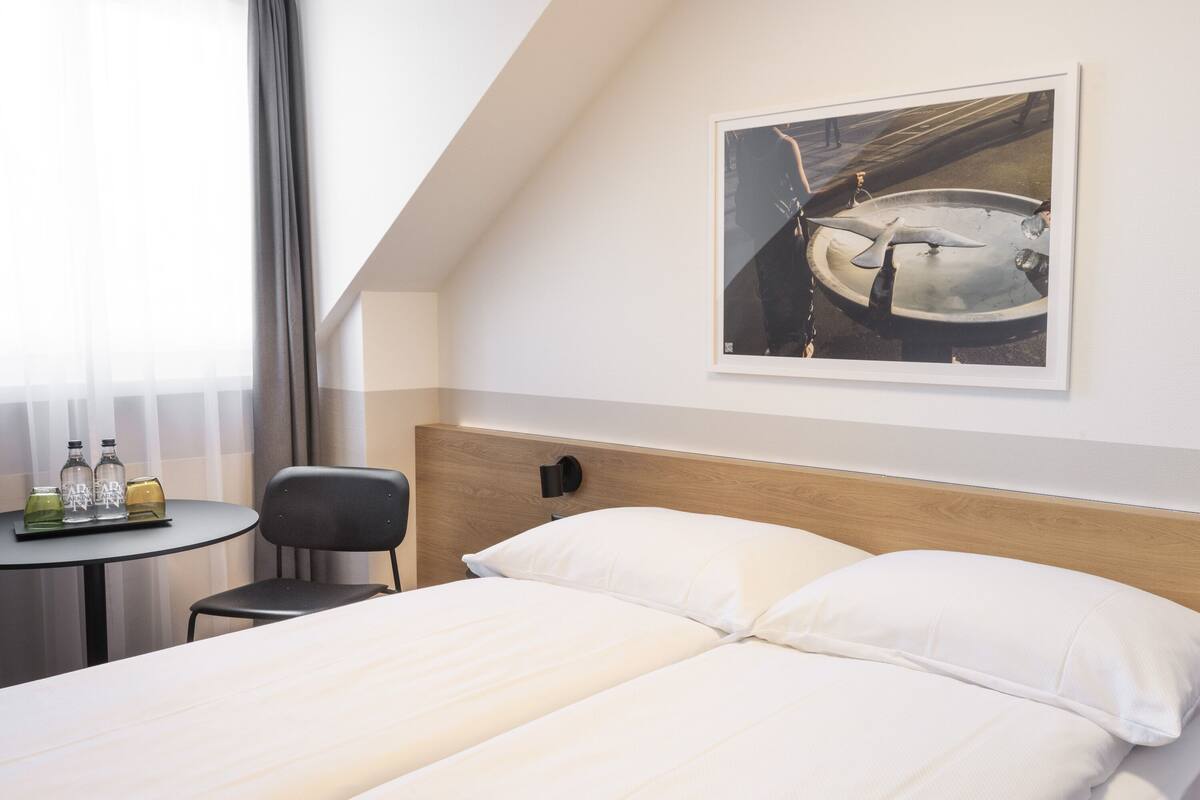 Ein Hotelzimmer mit einem Bett, einem kleinen Tisch mit Flaschen, einem Sessel und einem Bild an der Wand.