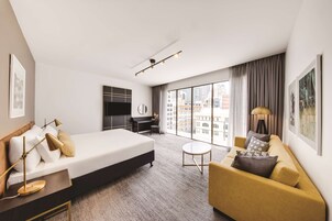 A modern hotel room with a large bed, a sofa, a desk, and a TV. The room has large windows with a city view.