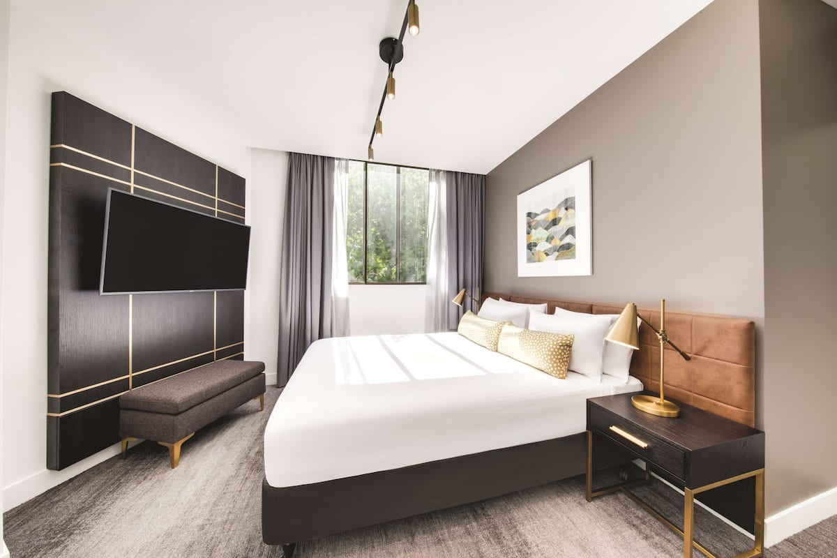 A modern bedroom with a large bed, a bench, a bedside table, a TV mounted on the wall, and a window with curtains.