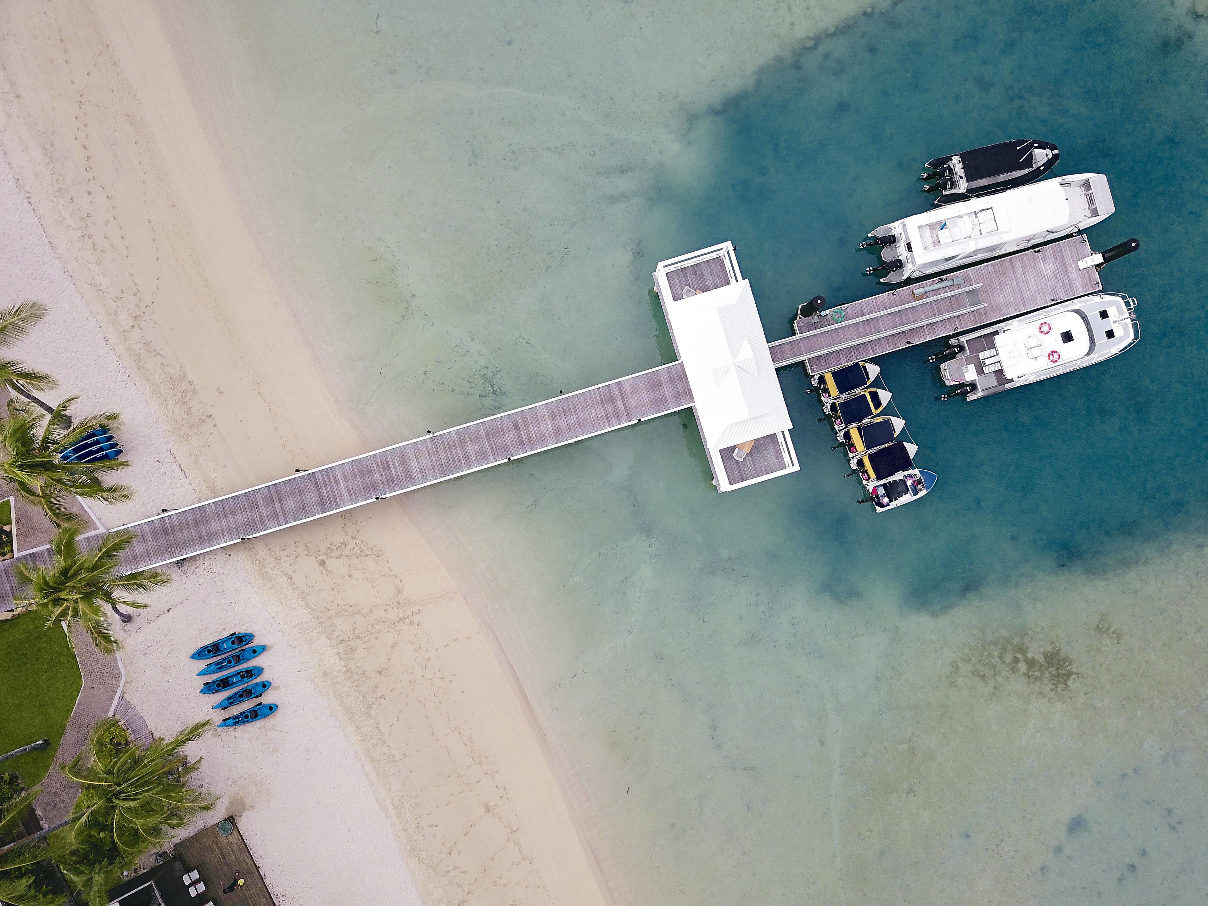 An aerial view of a beachside pier with boats docked, a walkway, and kayaks lined up on the sand.